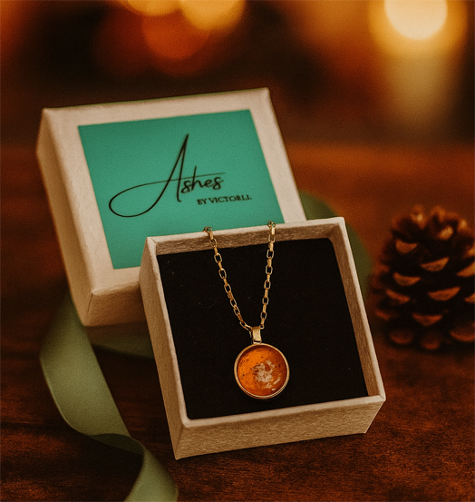 Gold memorial neclace with a yellow stone of cremation ash in glass in a white box with warm lighting and a winter theme of a pinecone and wooden table 